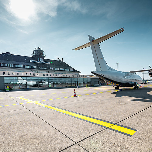 Vorfeld des Flughafens Braunschweig-Wolfsburg bei sonnigem Wetter. Rechts steht ein weißes Turboprop-Flugzeug, im Hintergrund sind das Terminal mit der Aufschrift „FLUGHAFEN BRAUNSCHWEIG-WOLFSBURG“ und der Kontrollturm zu sehen. Markierungen auf der Betonfläche führen durch das Bild, darüber spannt sich ein klarer blauer Himmel mit leichter Bewölkung.