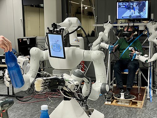 Ein Roboter greift im Vordergrund nach einer Flasche. Im Hintergrund sitzt ein Mensch in einer Vorrichtung und steuert den Roboter aus der Ferne.