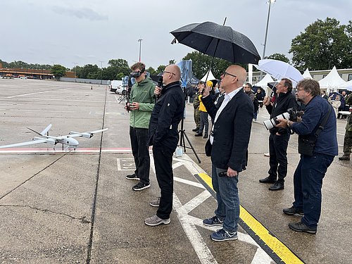 Mehrere Herren stehen neben einer Drohne, die wie ein Flugzeugt gestaltet ist. Zwei Herren haben Mikrophone in der Hand, um den Besuchenden Informationen &uuml;ber die Drohne zu vermitteln.