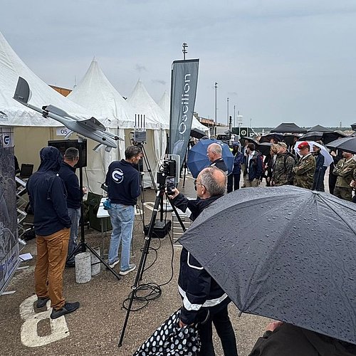 Eine Gruppe von Menschen mit aufgespannten Regenschirmen steht vor einem Zelt und h&ouml;rt einem Referenten zu. Am Zelt ist eine Drohne mit dem Namen Songbird sowie ein Ger&auml;t zur Identifizierung von Drohnen aufgestellt.