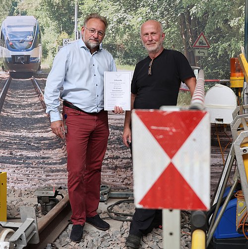 Zwei Personen stehen auf dem Übungsgleis in der Halle der CONDOR Technik und halten gemeinsam eine Urkunde zum zehnjährigen Dienstjubiläum. Rund um das Gleis sind verschiedene Geräte der Gleisbaustellensicherung aufgebaut, darunter Warnleuchten, eine rot-weiße Signaltafel und weitere technische Anlagen. Im Hintergrund zeigt eine großformatige Fotowand einen Zug auf einer von Bäumen gesäumten Bahnstrecke.
