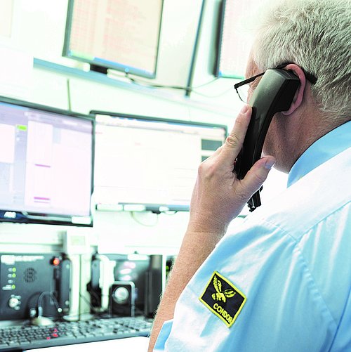Ein Mann sitzt mit einem Telefonhörer in der Hand vor mehreren Bildschirmen. Auf dem Hemd befindet sich das Logo der Firma CONDOR Schutz- und Sicherheitsdienst.