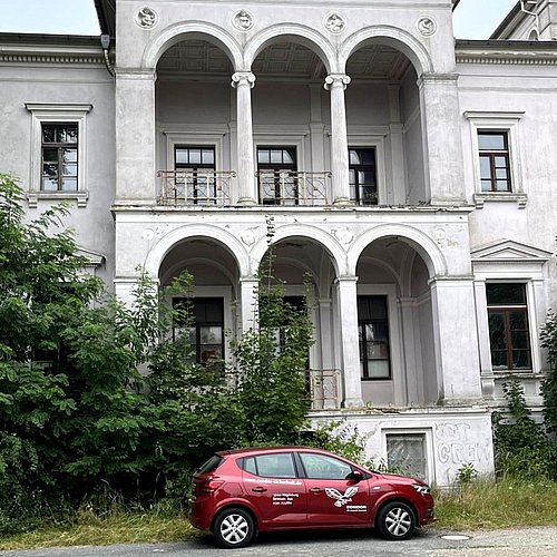 Vor einem Gebäude steht ein rotes Auto der Firma Condor Schutz und Sicherheit.