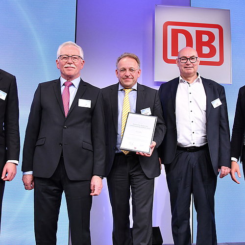 DB-Lieferantenprädikat für Kombiausbildung Fachkraft für Gleisbausicherung Fünf Personen stehen nebeneinander. Die Person in der Mitte hält eine Urkunde in der Hand. Im Hintergrund befindet sich das LOGO der Deutschen bahn.