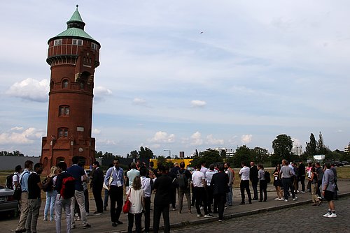 Eine Menschengruppe schaut in den Himmel und beobachtet eine Drohne, die auf ein Gebäude zu einem Gebäude fliegt.
