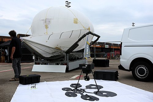 Im Vordergrund des Bildes befindet sich eine Drohne, die auf einer Plane mit dem Logo der Drone it GmbH steh. Im Hintergrund befindet sich ein mannshoher und mit Helium gef&uuml;llter Balon, der Heli-Kite genannt wird.