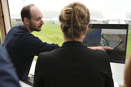 Ein Mann erklärt einer Frau den Ablauf eines Drohnenfluges, indem er auf eine Karte zeigt, die auf einem Bildschirm angezeigt wird.