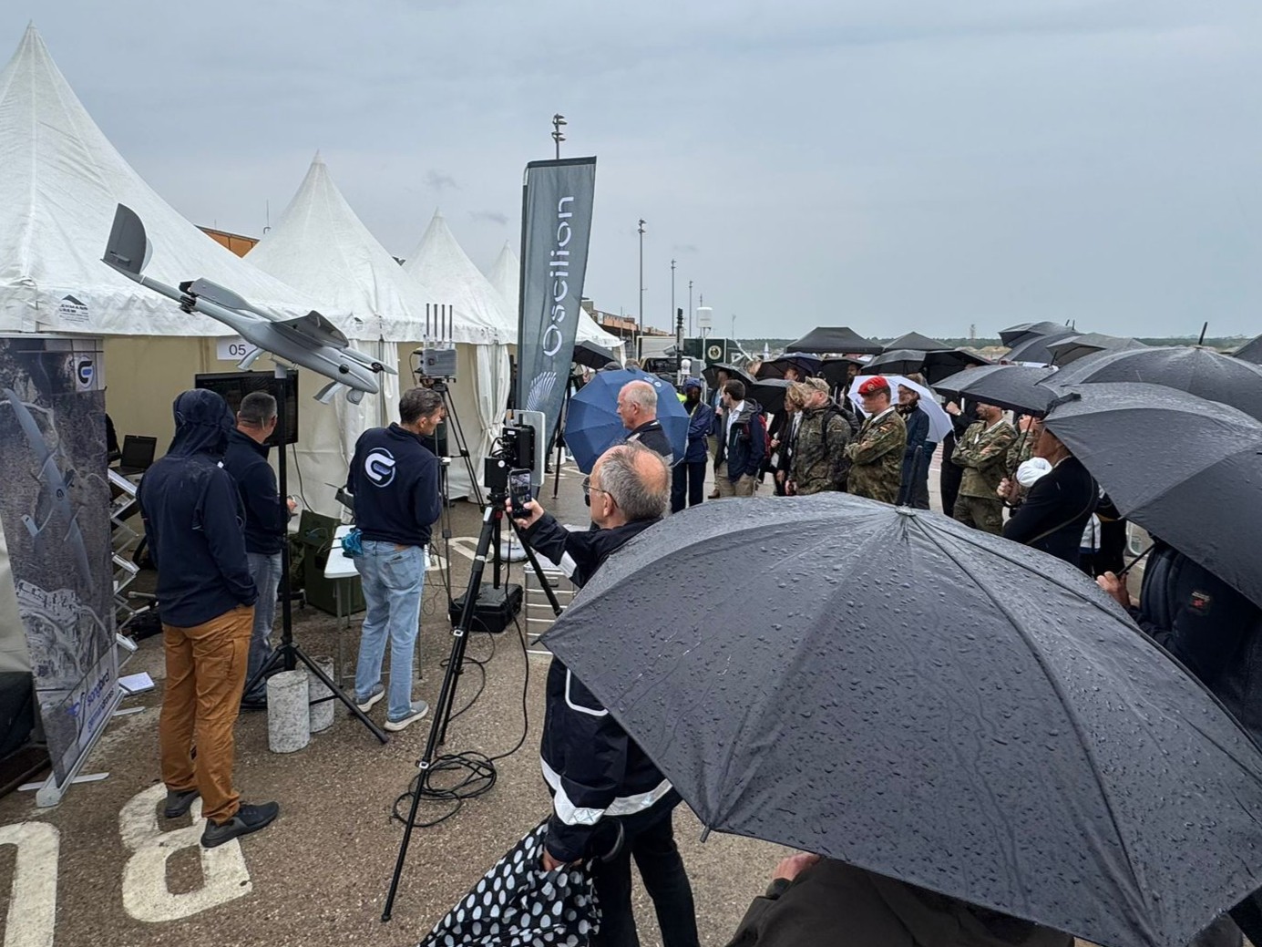 Eine Gruppe von Menschen mit aufgespannten Regenschirmen steht vor einem Zelt und h&ouml;rt einem Referenten zu. Am Zelt ist eine Drohne mit dem Namen Songbird sowie ein Ger&auml;t zur Identifizierung von Drohnen aufgestellt.