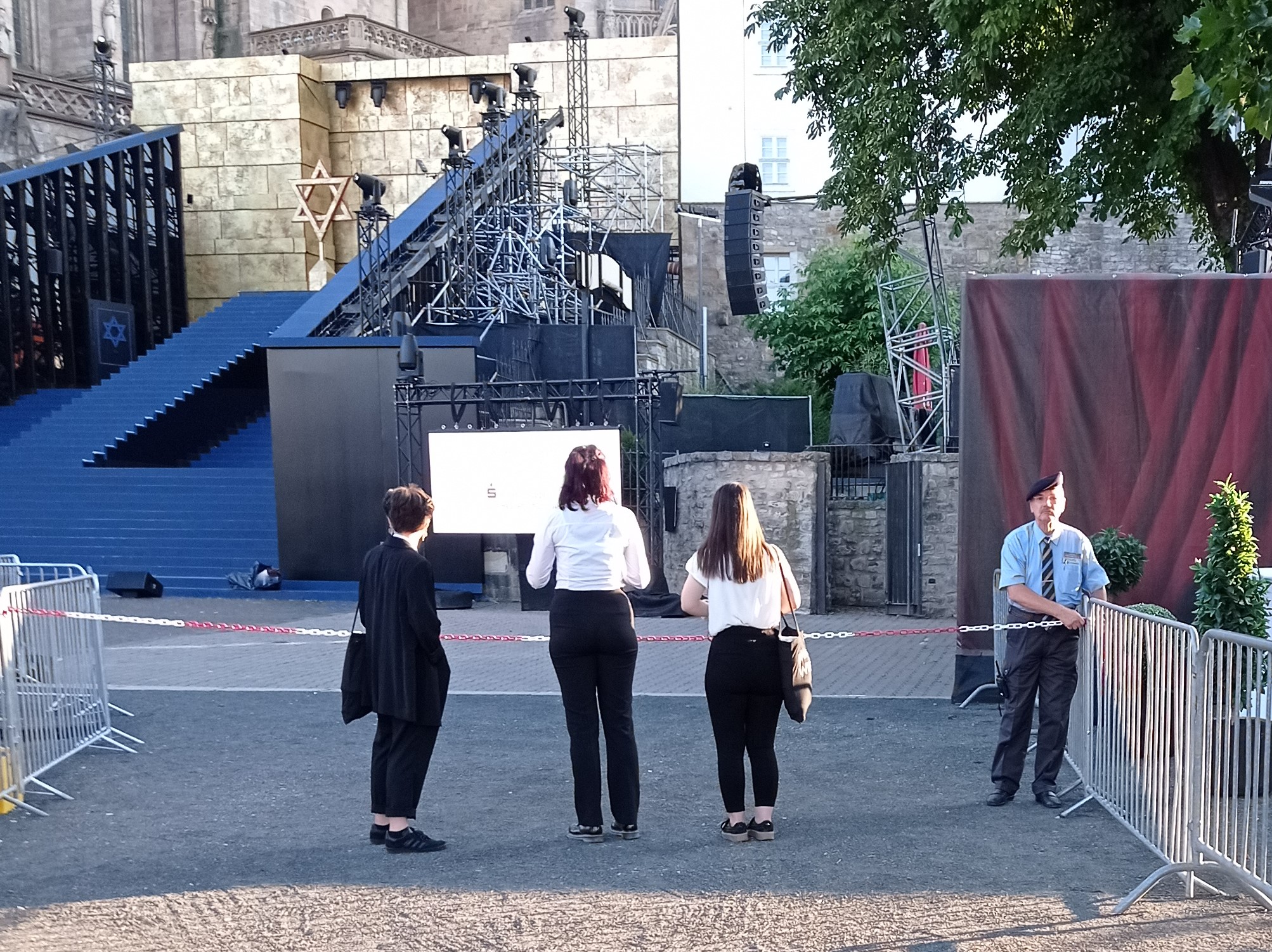 Drei Frauen stehen vor einem Absperrband und schauen auf die Bühne der DomStufen-Festspiele. Ein Mann sichert die Absperrung.